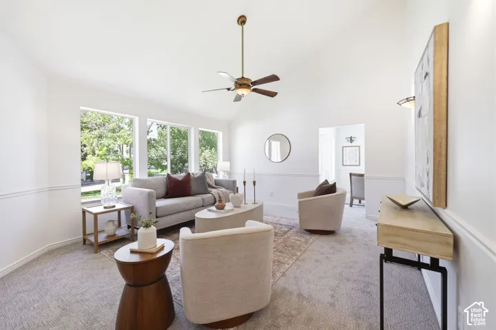 Bright, open-concept living space with triple glass doors framing natural light, ceiling fan with bronze finish, curved upholstered seating, wood-finish media console, round wood accent table, and neutral carpet flooring throughout