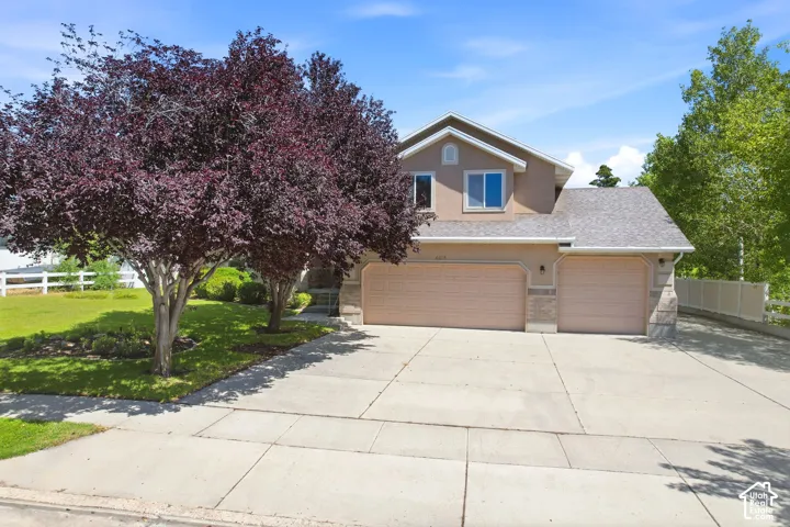 Tan two-story home with a three-car garage, concrete driveway, and mature landscaping including a burgundy-leafed tree and established greenery