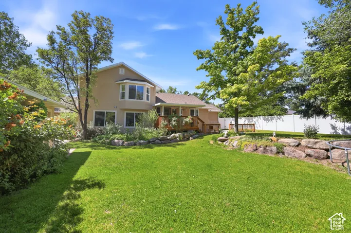 Expansive manicured lawn with mature shade trees and tiered landscape beds, backed by a two-story residence featuring a tan exterior, multi-pane windows, and an attached deck with wooden railings overlooking the grounds