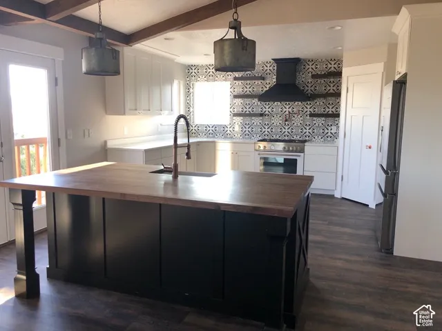 Kitchen with wood-finish flooring, exposed wood beams, a central island with a wood countertop and integrated sink, a tiled backsplash, and stainless steel appliances