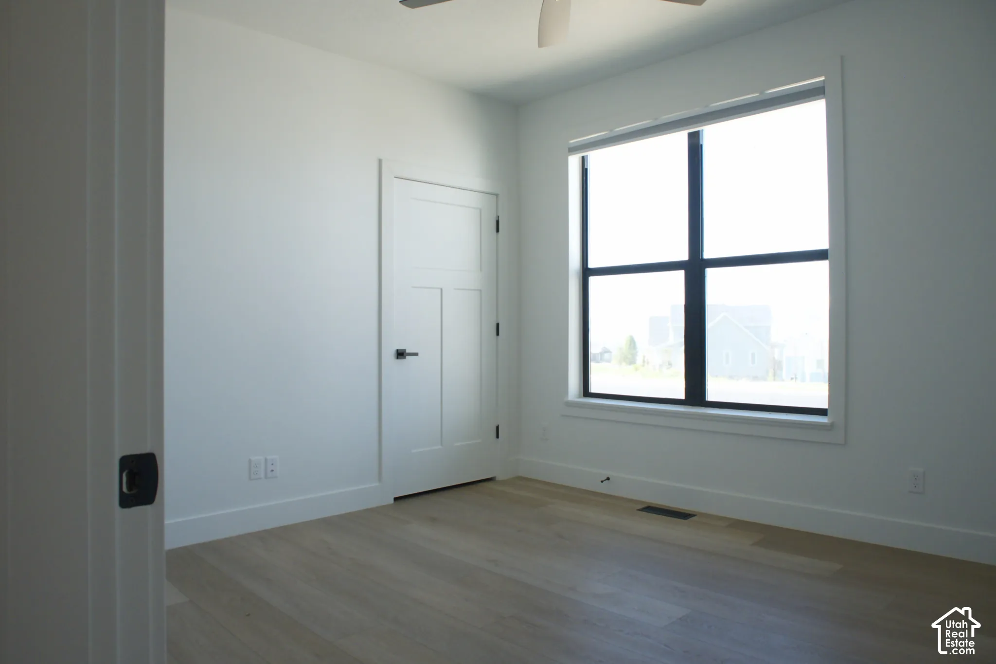 White walls and ceiling, wood-finish flooring, white solid core door with black hardware, large black-framed window, and a ceiling fan
