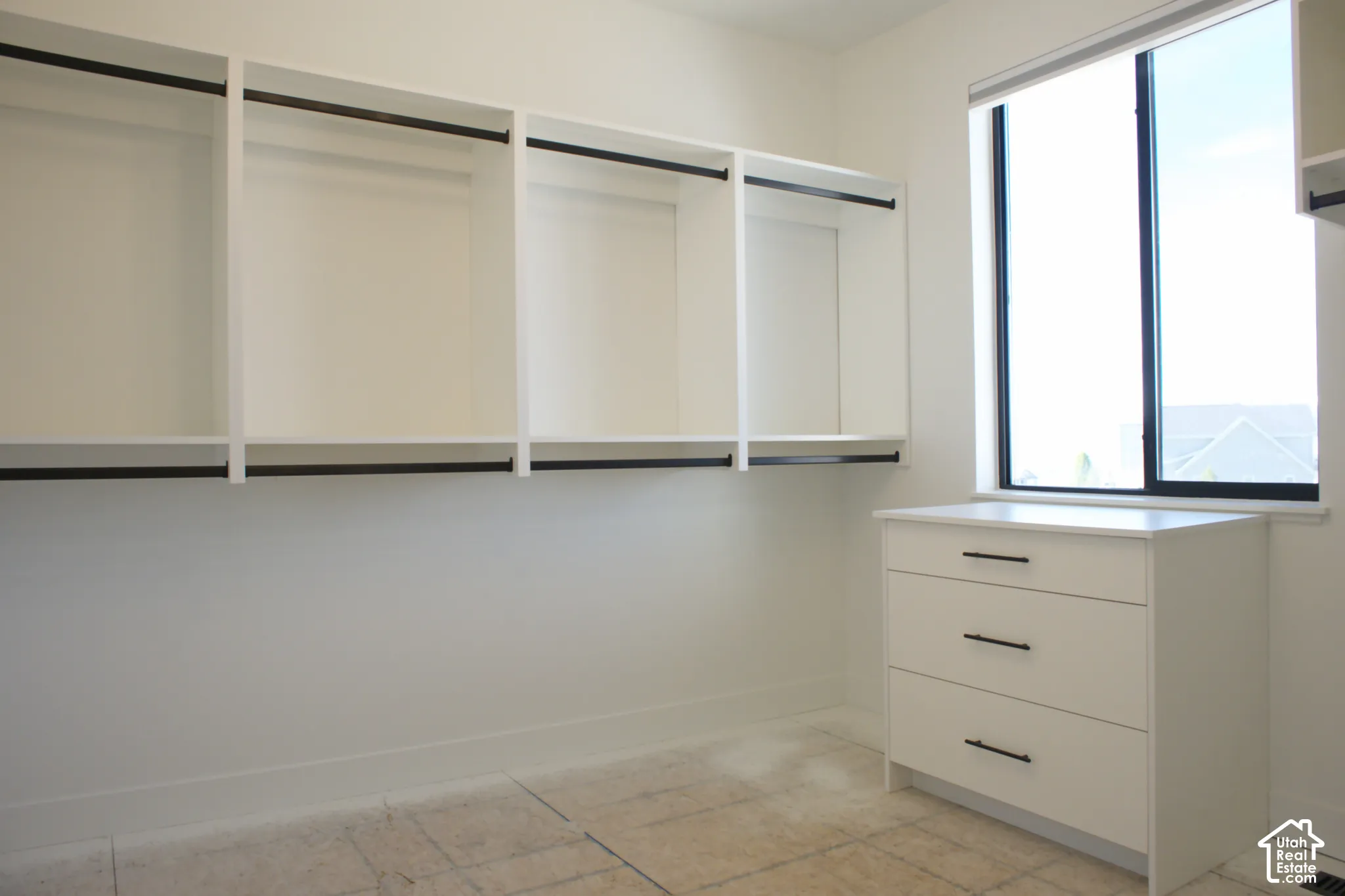 Built-in white shelving with black hanging rods, white three-drawer dresser with black pull handles, and a large window