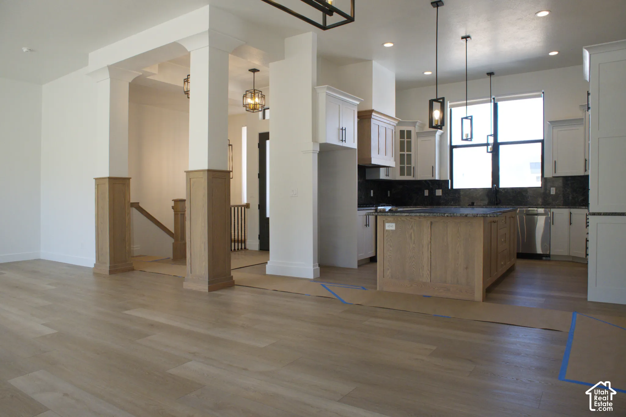 Open-concept living and kitchen area featuring wood-finish flooring throughout, a large island with dark countertop, white cabinetry, stainless steel appliances, pendant lighting, and architectural columns defining the space