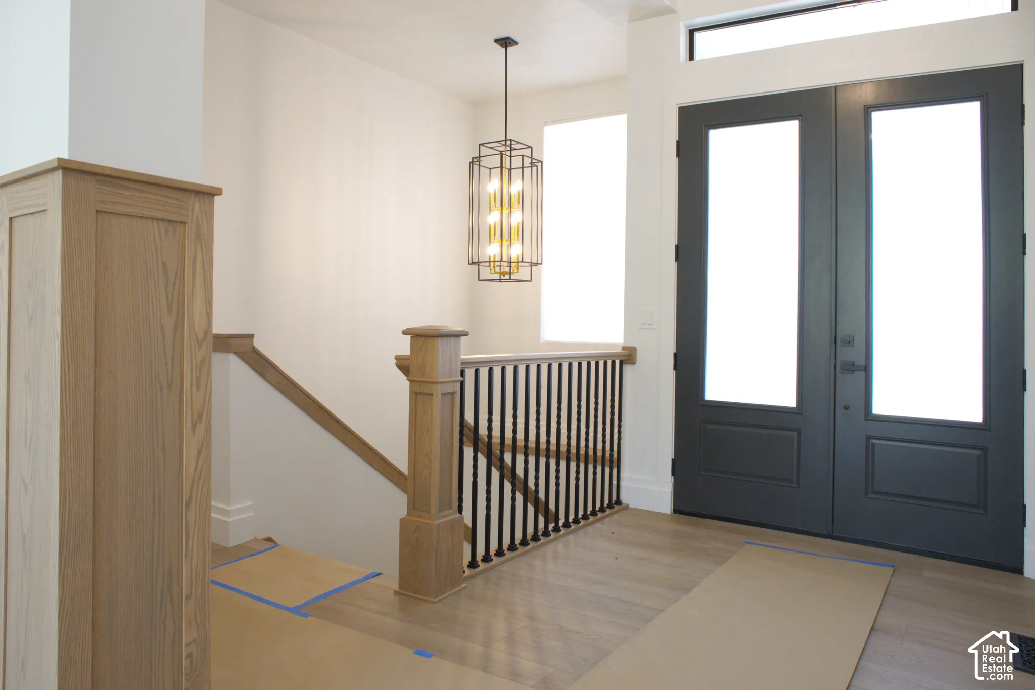 Light wood-finish flooring, dual entry doors, transom window, contemporary chandelier, and wood staircase with iron balusters