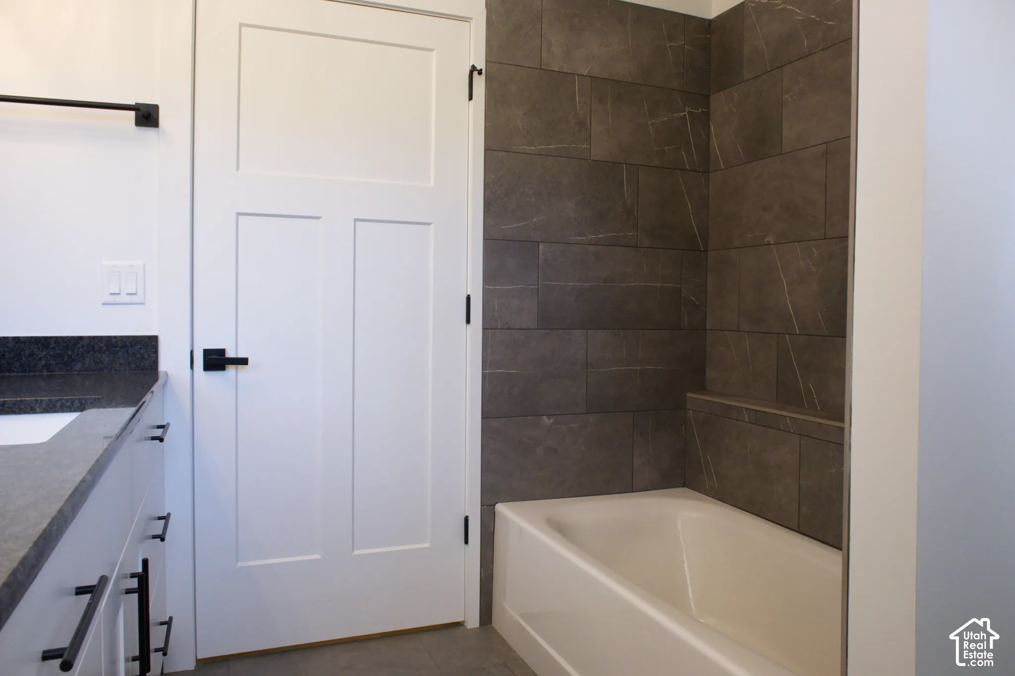 Bathroom featuring a white vanity with a dark countertop, white flat-panel door with black hardware, and a tub with dark tile surround and built-in ledge