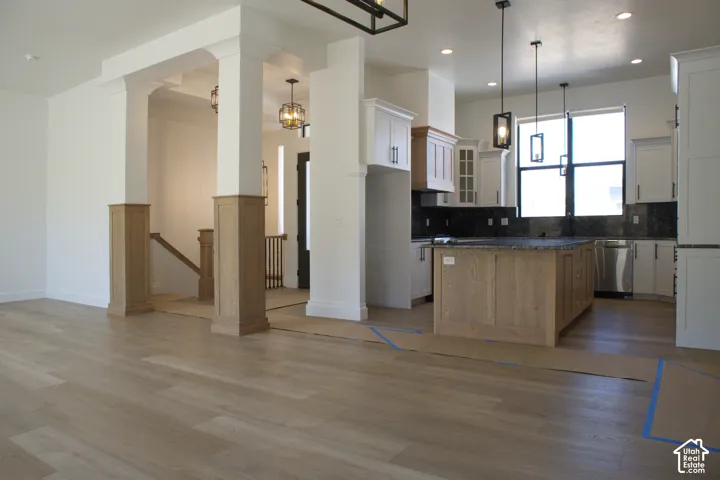 Open-concept living and kitchen area featuring wood-finish flooring throughout, a large island with dark countertop, white cabinetry, stainless steel appliances, pendant lighting, and architectural columns defining the space