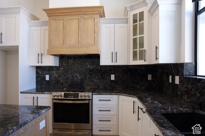 White shaker-style cabinetry with matte black hardware, dark natural stone countertops, a light wood-finish range hood, and a stainless steel oven