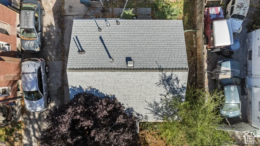 Asphalt shingle roof featuring multiple metal roof vents and chimney flues, framed by established mature trees and perimeter wood fencing