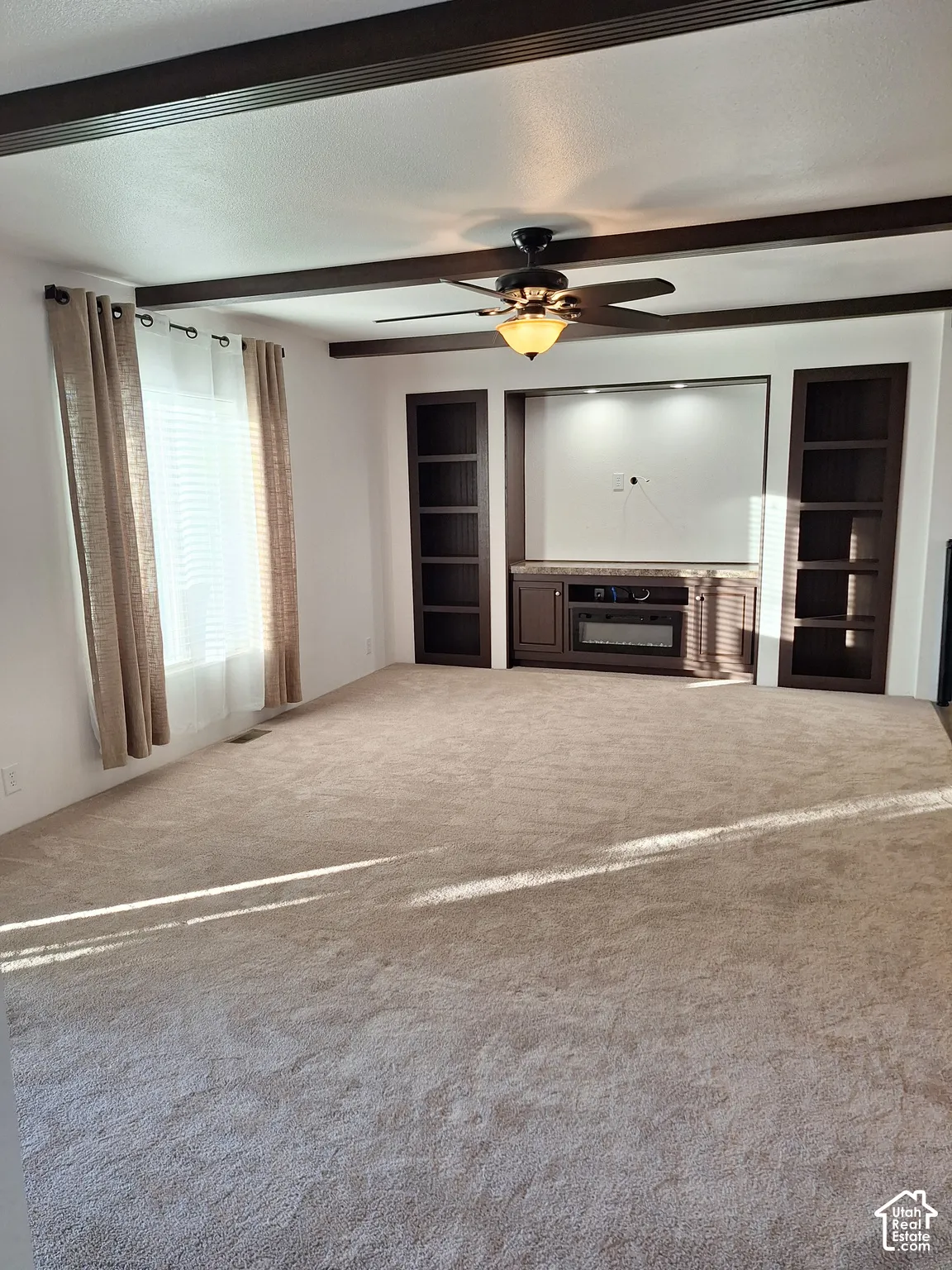 Recessed entertainment center with built-in shelving, decorative ceiling beams, ceiling fan with integrated lighting, and neutral-toned carpeting