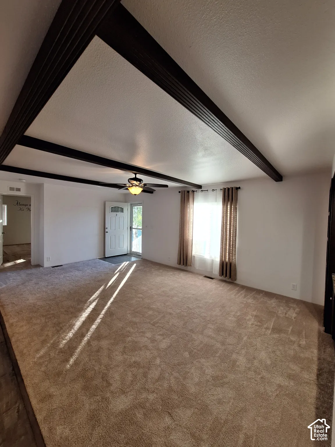 Vaulted ceiling with dark wood-finish beams, ceiling fan with integrated lighting, broad window, and plush carpet flooring
