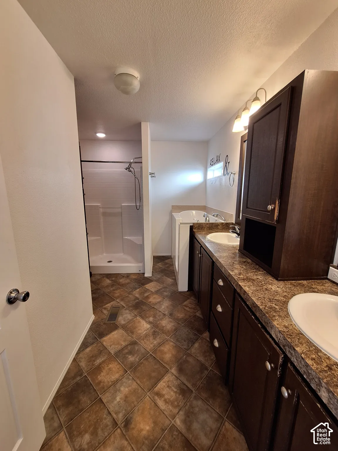 Dual vanity with dark wood-finish cabinetry, a drop-in soaking tub, a stand-up shower, and patterned floor tiling