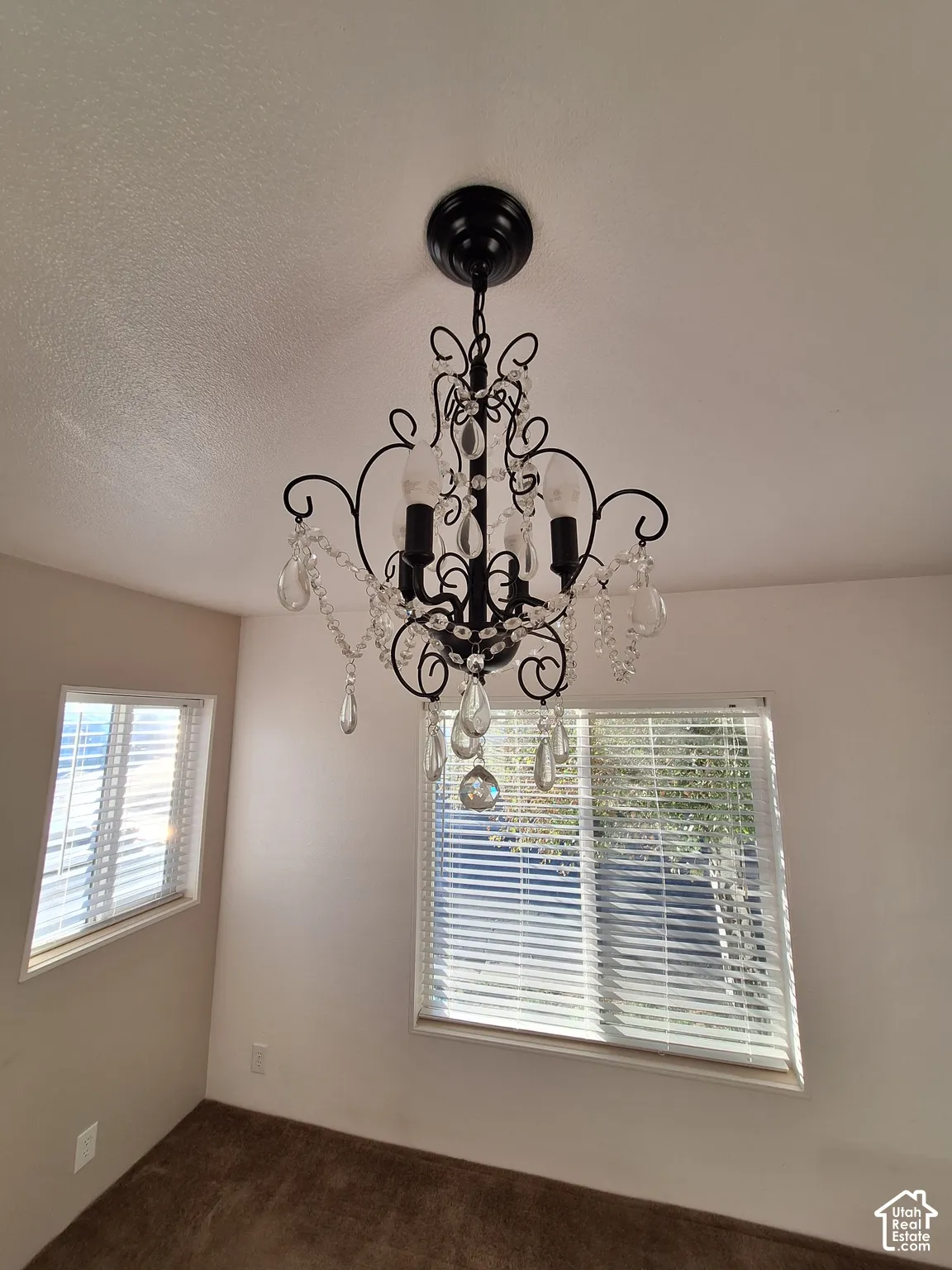 Black wrought iron chandelier with clear crystal accents, two windows with white blinds, neutral wall paint, and brown carpet