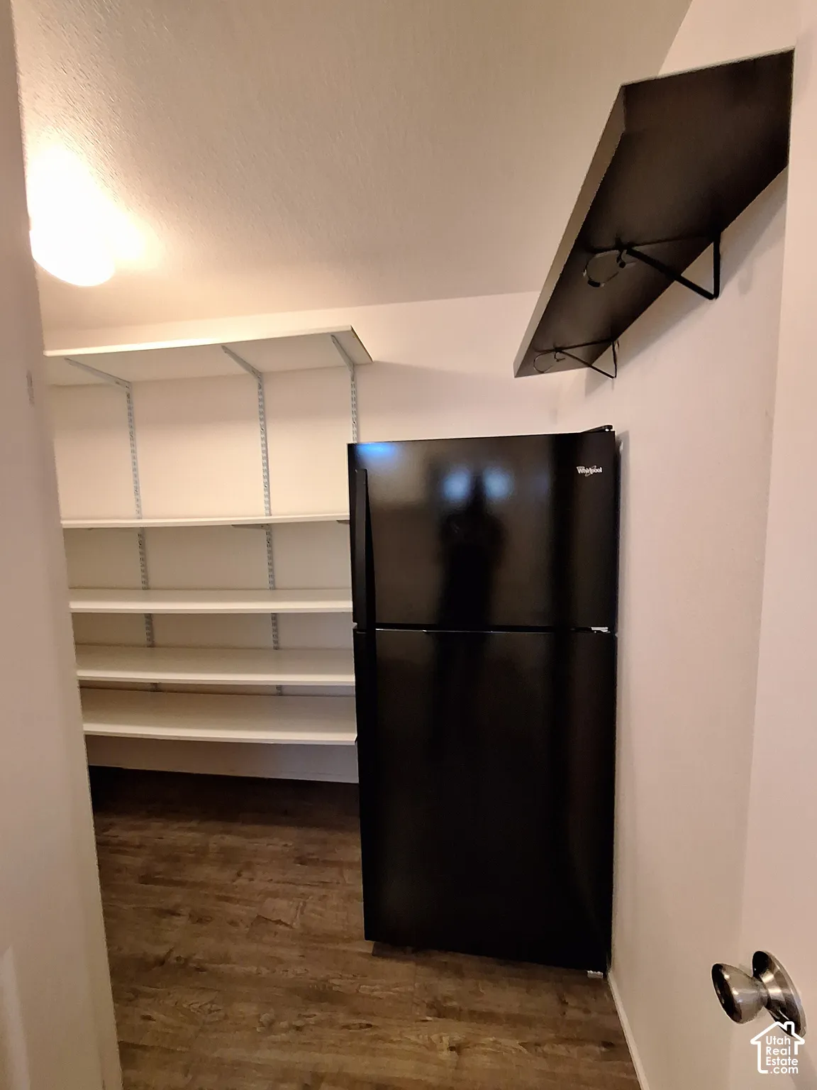 Compact utility space featuring white shelving with multiple tiers for storage, wood-finish flooring, black refrigerator, recessed ceiling lighting, and mounted exhaust hood