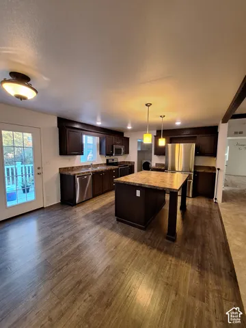 Kitchen with wood-finish flooring, dark wood cabinetry, stainless steel appliances, and an island with a stone-finish countertop
