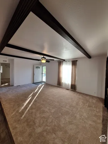 Vaulted ceiling with dark wood-finish beams, ceiling fan with integrated lighting, broad window, and plush carpet flooring