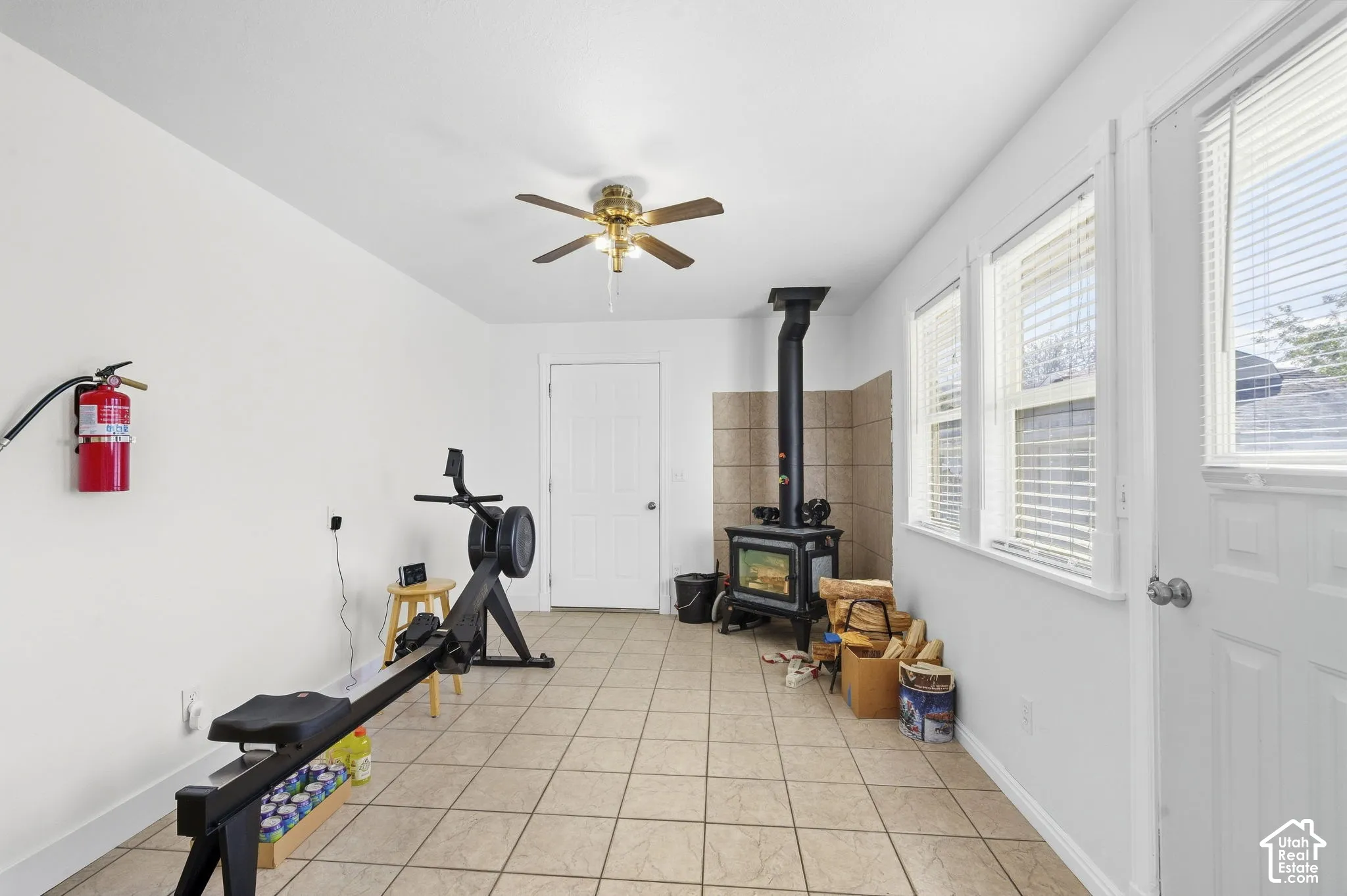 Room featuring tile flooring, a ceiling fan, multiple windows with blinds, a white paneled door, and a wood-burning stove with a tiled surround