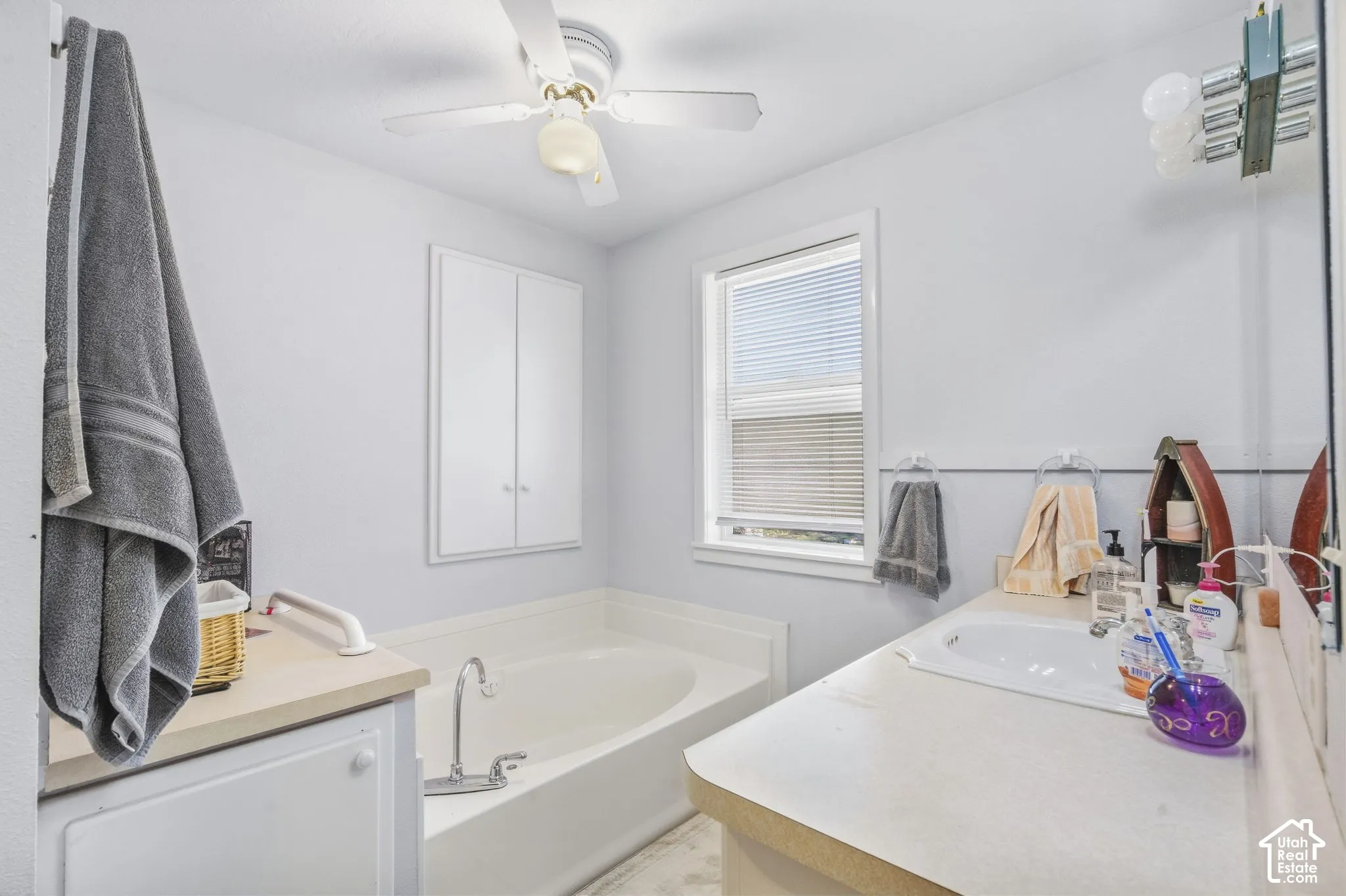 Integrated soaking tub, built-in wall cabinet, single-basin vanity with ample counter space, window with blinds, and a ceiling fan with light fixture