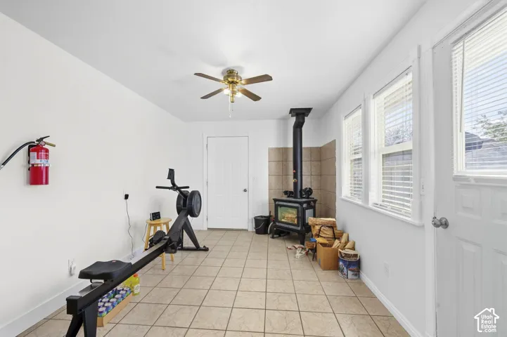 Room featuring tile flooring, a ceiling fan, multiple windows with blinds, a white paneled door, and a wood-burning stove with a tiled surround