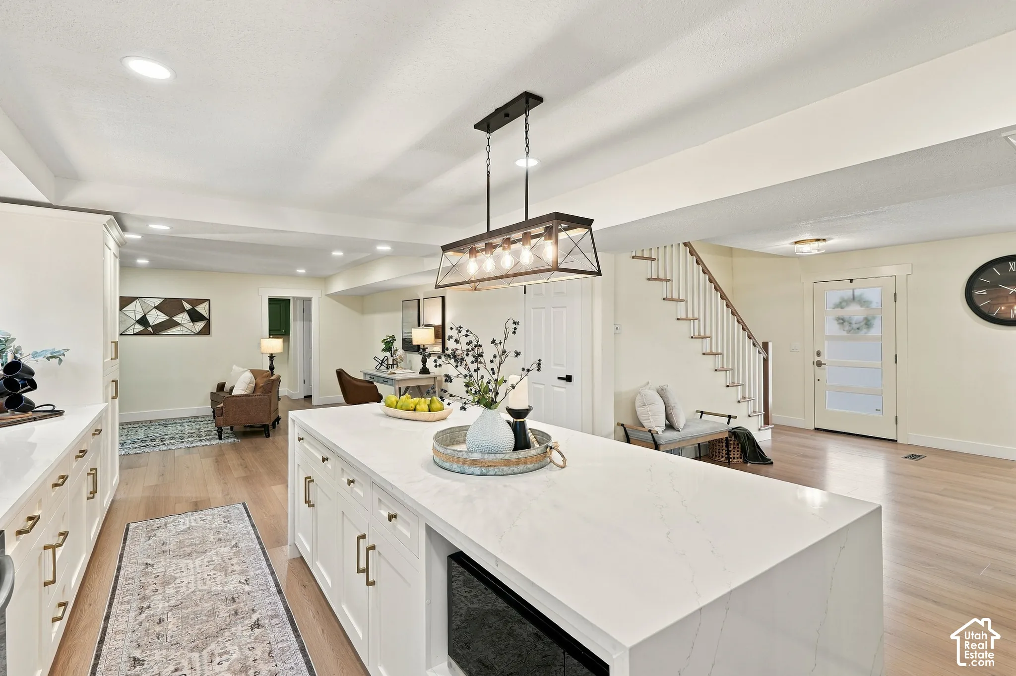 White kitchen island with a waterfall countertop, white cabinetry with gold-toned hardware, wood-finish flooring, recessed lighting, and an open staircase with white balusters