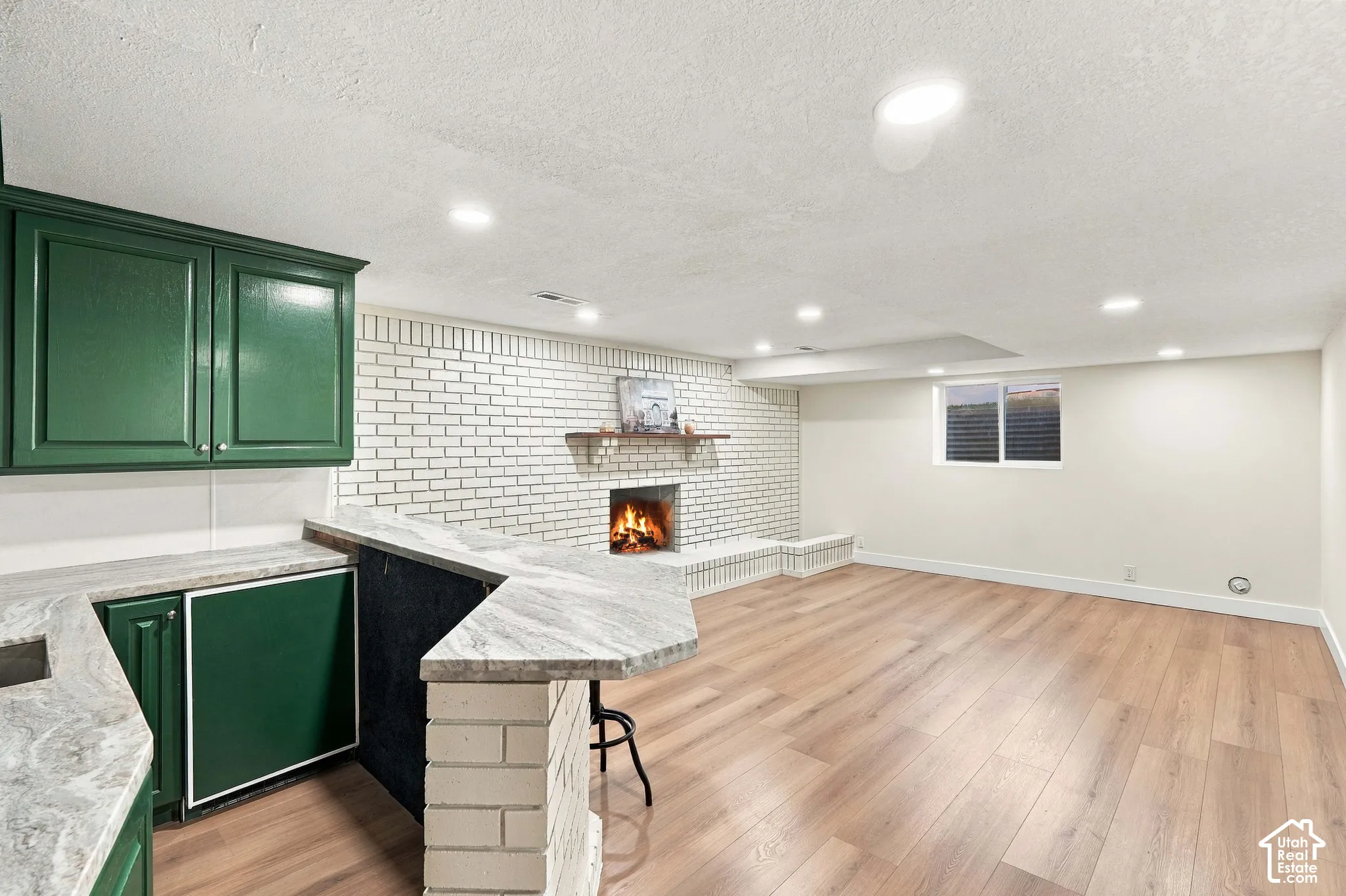 Wet bar with green cabinetry and stone-finish countertops, brick-finish fireplace with wood mantel, wood-finish flooring, and recessed lighting