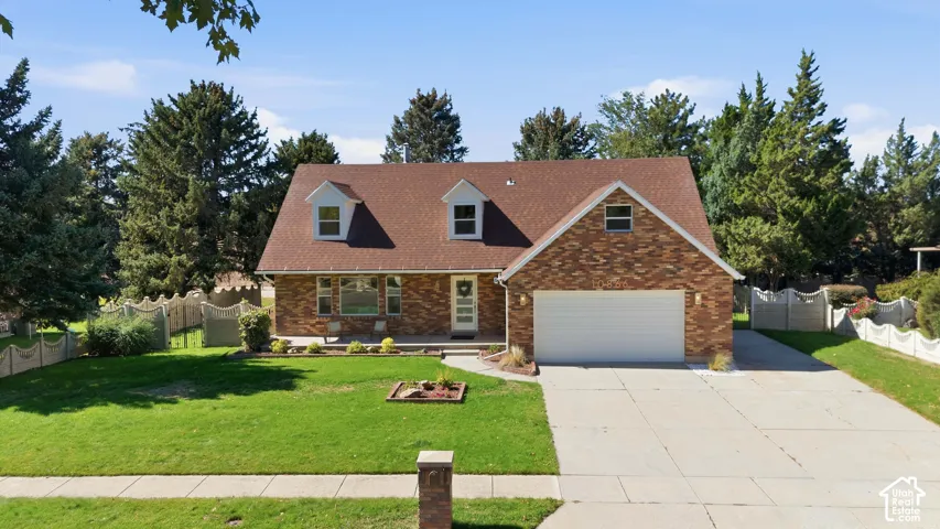 Brick exterior with dormer windows, attached garage, concrete driveway, and mature landscaping