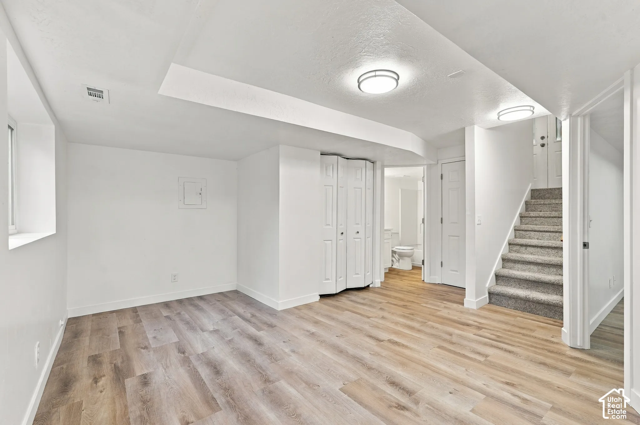 Spacious room featuring wood-finish flooring, white walls, flush mount lighting, and a carpeted staircase