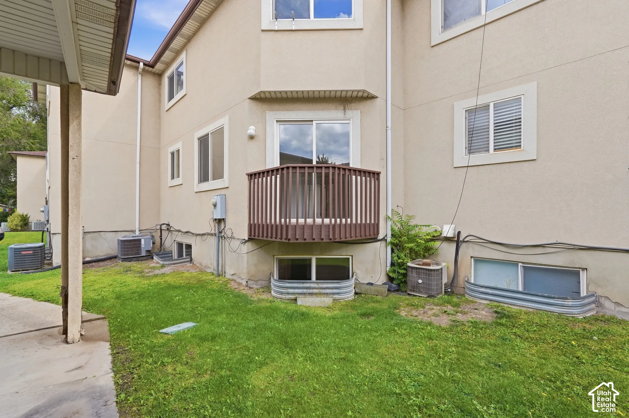 Exterior stucco finish with a ground-level patio, upper-level balcony with wood railing, and multiple window wells