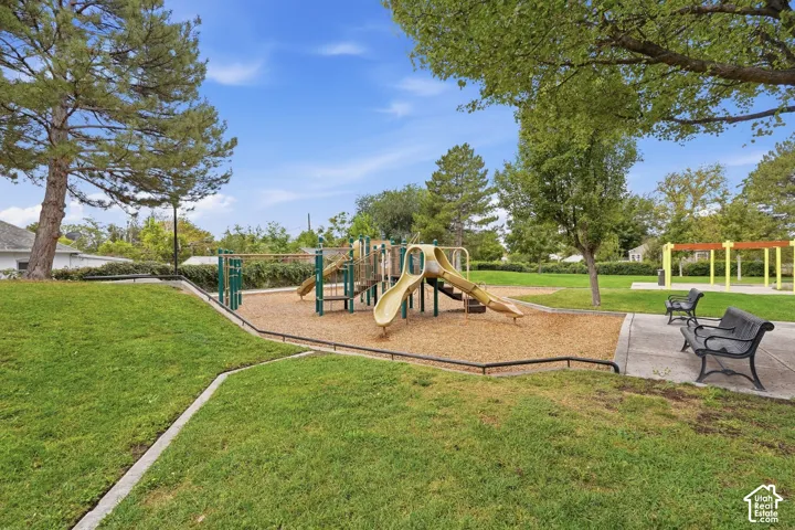 Expansive park area featuring a multi-level playground structure, two park benches, a large grassy expanse, and mature trees