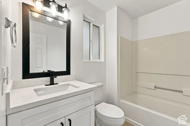 Bathroom featuring a modern vanity with a white countertop and integrated sink, a rectangular mirror with a black frame, a wall-mounted light fixture, a toilet, and a bathtub with a shower surround