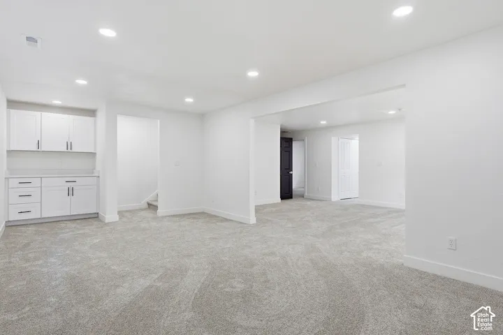 Recessed lighting illuminates the open-concept space, featuring plush, gray carpeting, a built-in wet bar with white cabinetry, and a countertop
