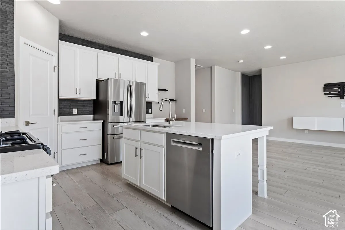 White shaker cabinetry with stainless steel appliances, quartz countertops, a kitchen island with a sink, and wood-finish flooring