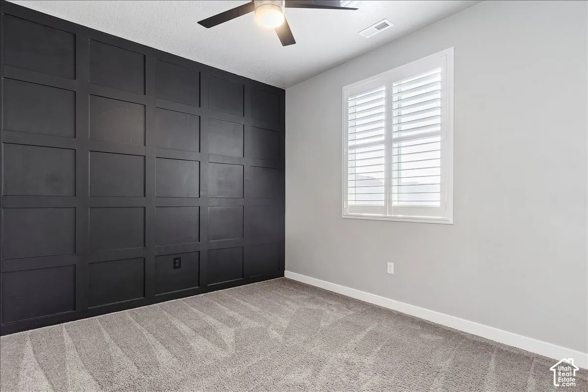 Accent wall with panel detailing, light-colored walls, window with plantation shutters, ceiling fan with integrated lighting, and carpeted flooring