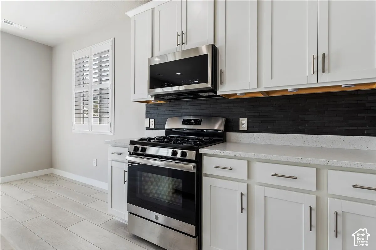 White shaker-style cabinetry with brushed nickel hardware, stainless steel gas range with overhead microwave, dark tile backsplash, light-colored countertops, and wood-finish flooring