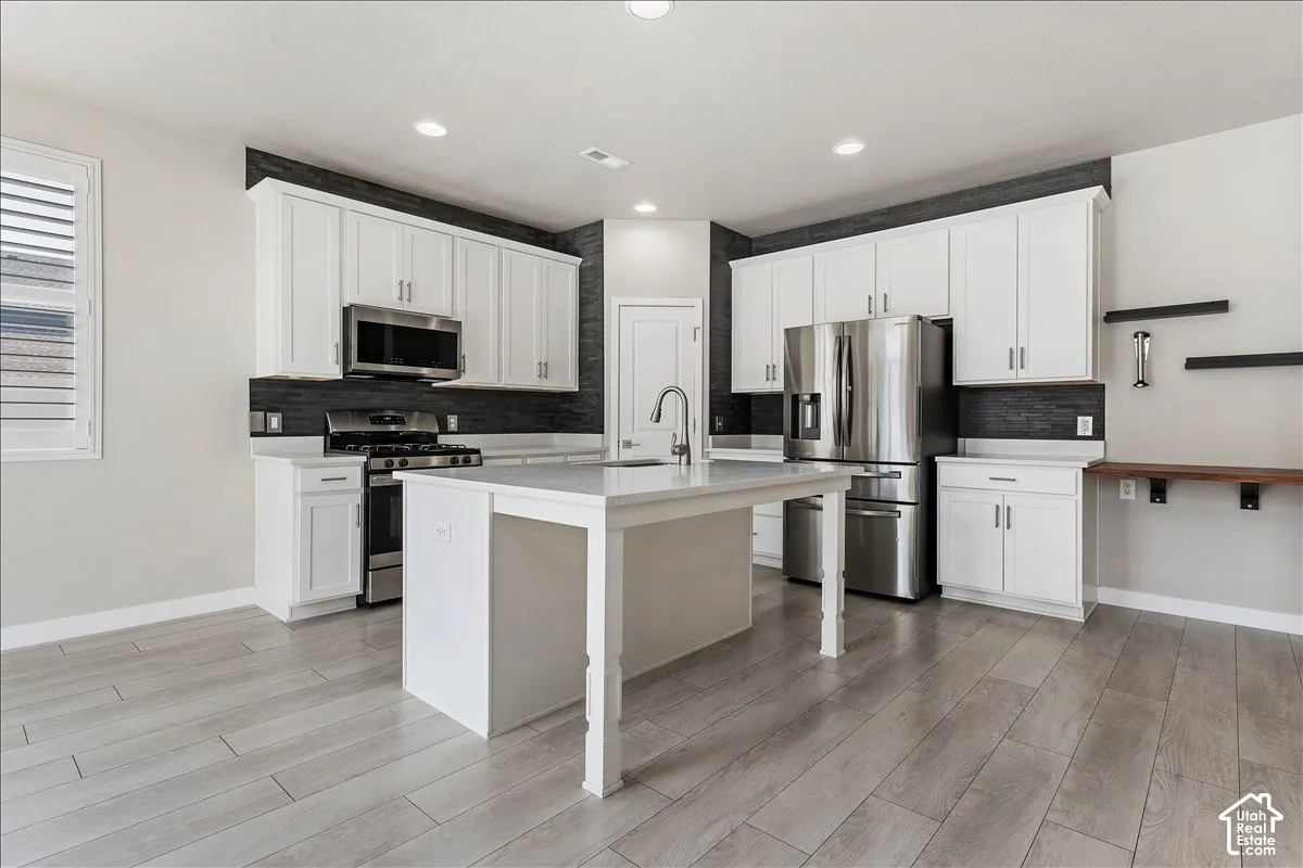 Kitchen featuring an island, stainless steel appliances, wood-finish flooring, white cabinetry, and a dark tile backsplash