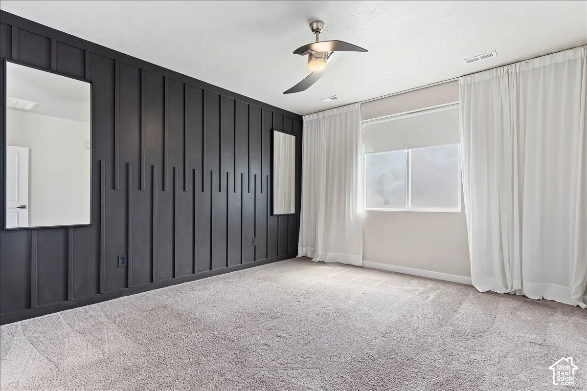 Accent wall with textured paneling, light gray carpeting, ceiling fan with integrated lighting, and window with roller shade and drapes