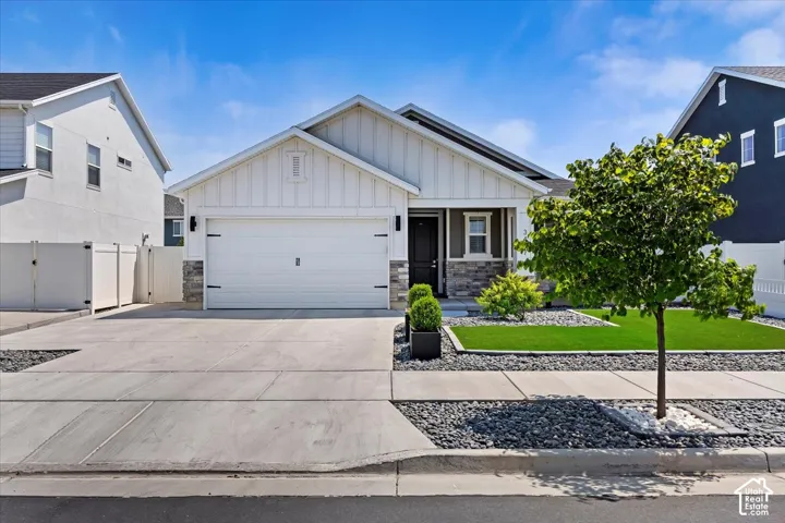 White horizontal siding with board and batten accents, attached garage, stone accents on the facade, concrete driveway, and a manicured front lawn with decorative rock landscaping