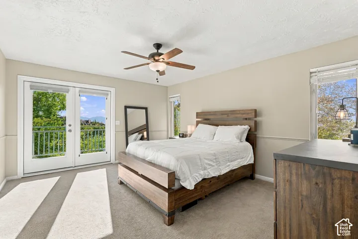 Primary bedroom suite featuring a wood-finish bed frame, ceiling fan with integrated lighting, carpet flooring, and dual glass doors opening to a private balcony with scenic outdoor vistas