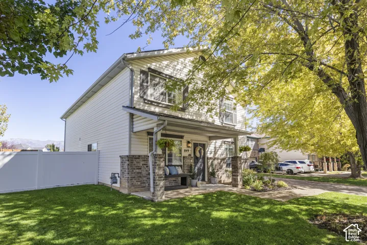 Two-story residence featuring white siding, a covered front porch with stone pillars, a front lawn with mature trees, and a white privacy fence