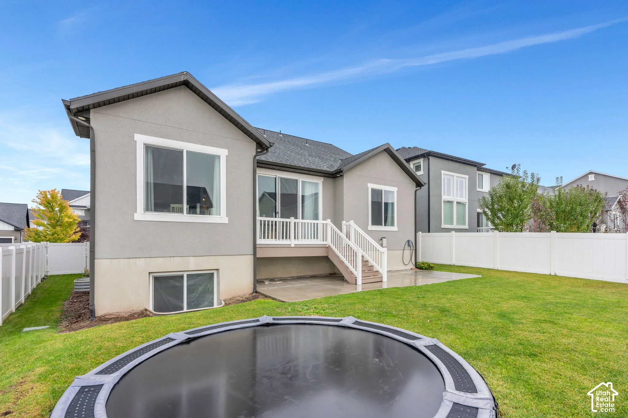 Stucco exterior with white trim, a deck with white railings and stairs, a concrete patio, a manicured lawn, and a white vinyl fence