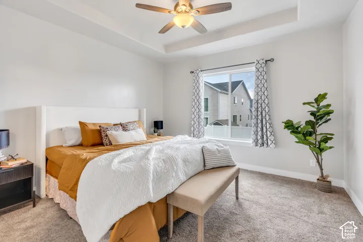 Tray ceiling, ceiling fan with integrated lighting, large window with white trim, and light-toned carpeting