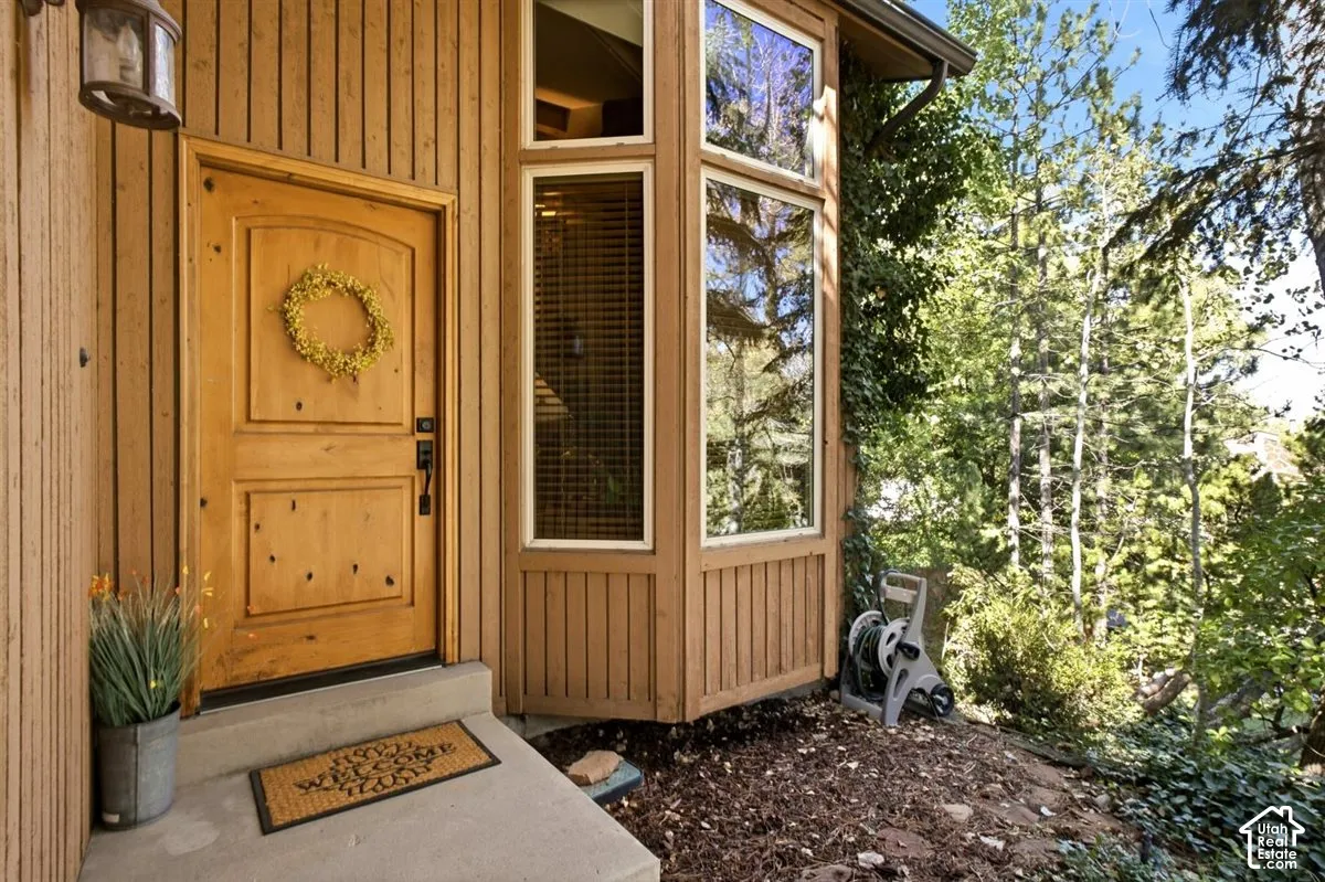 Vertical wood siding with a natural finish, wood panel door with arch detail, bay window with multiple panes, concrete steps, and surrounding mature trees