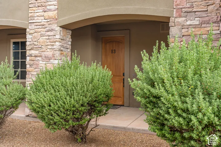 Stucco exterior with stone veneer accents, natural wood-finish entry door, covered entry, concrete walkway, and gravel landscaping with established shrubs