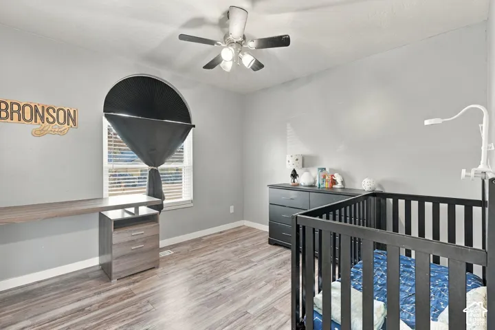 Wood-finish flooring, ceiling fan with integrated lighting, arched window with horizontal blinds, built-in desk with storage drawers, and gray wall finish