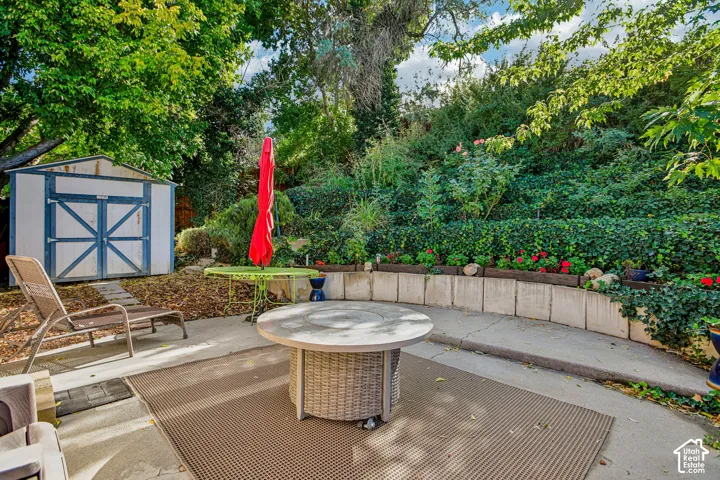 Paved patio with a wicker-style table, concrete retaining wall, a variety of mature plantings, and an accessory storage shed