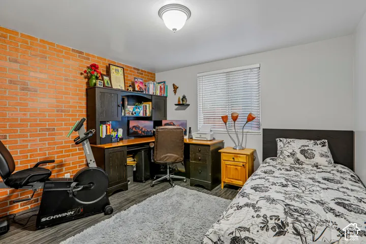 Exposed brick accent wall, wood-finish flooring, built-in desk with hutch, window with blinds, and recessed ceiling light fixture