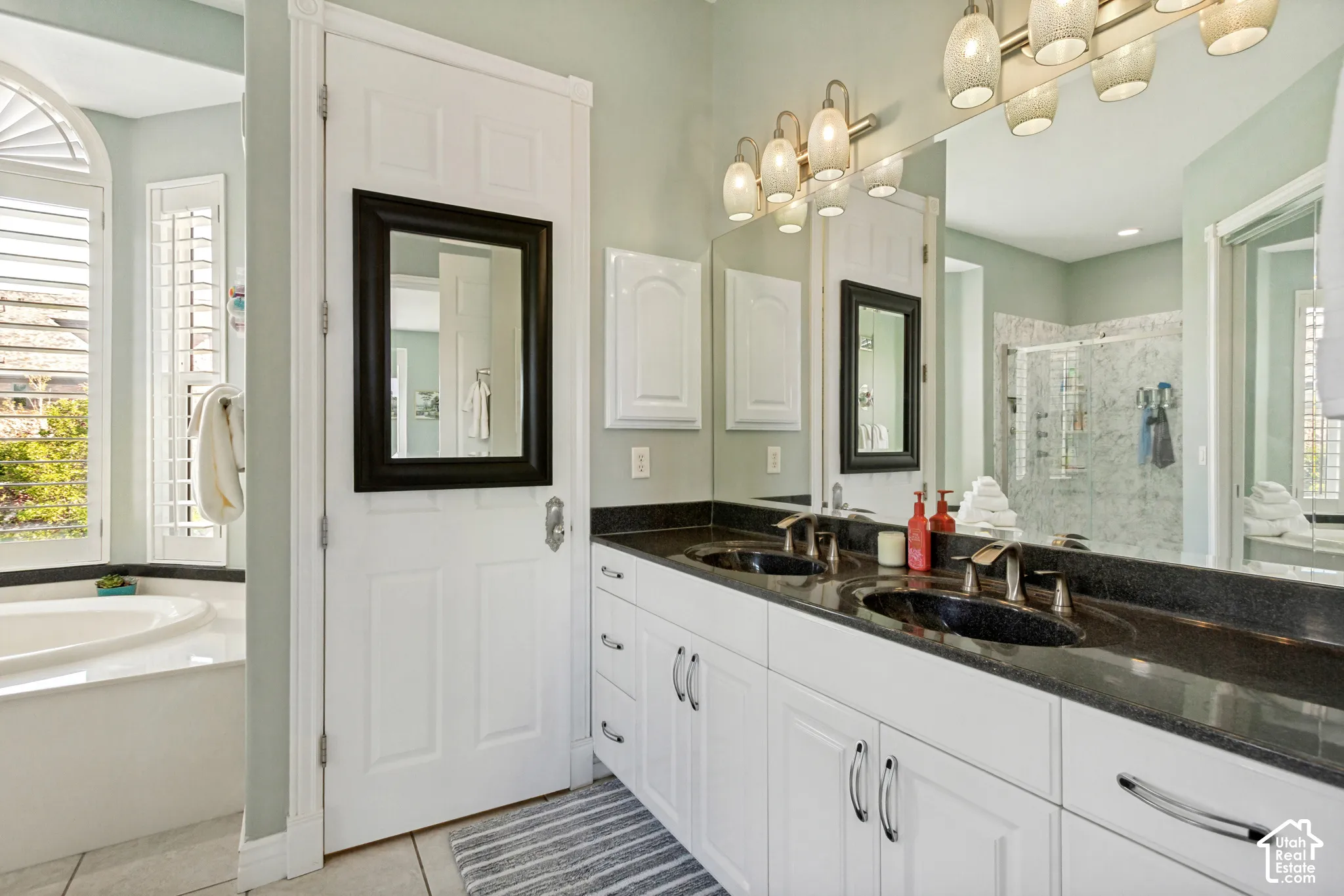 Dual vanity with dark countertop and undermount sinks, white cabinetry with chrome hardware, full-width mirror, and multi-bulb light fixture