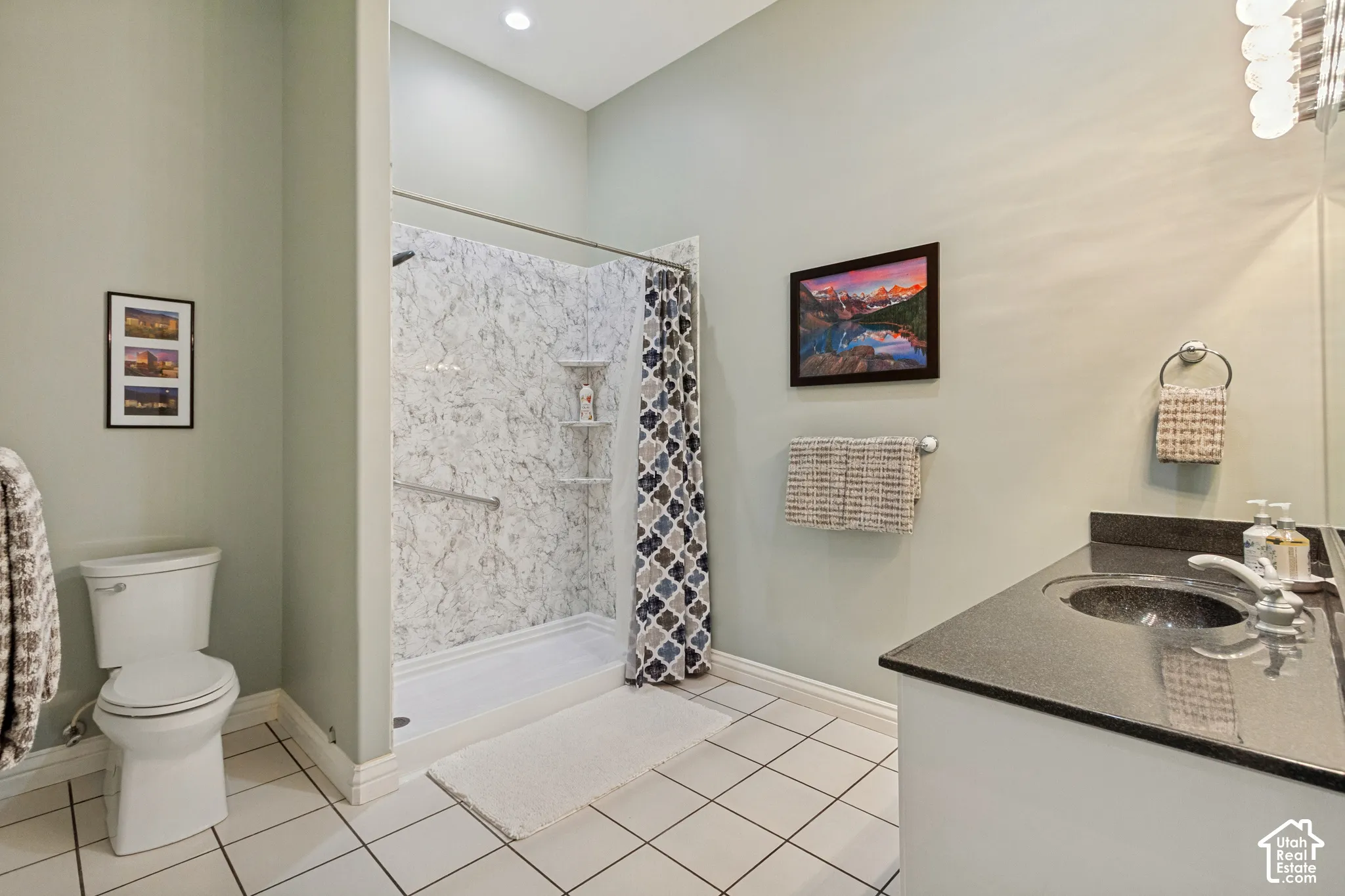 Tiled flooring, white toilet, shower with patterned wall surround, vanity with dark countertop and integrated sink, recessed lighting