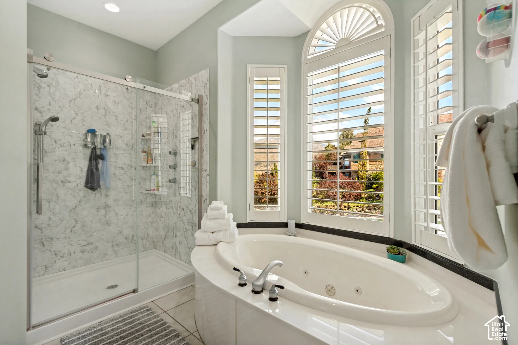 Soaking tub with integrated jets, arched window with plantation shutters, walk-in shower with glass enclosure, white marble-patterned shower surround, and dual shower heads