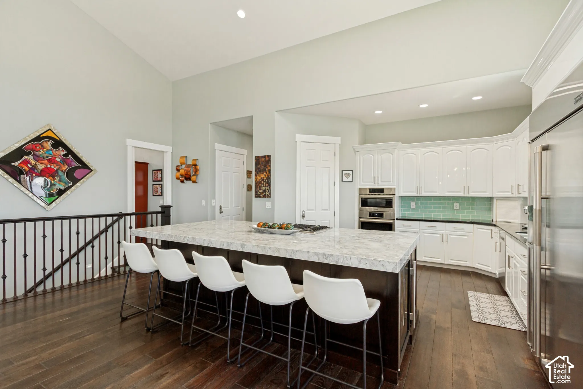 Vaulted ceiling with recessed lighting, dark wood-finish flooring, white cabinetry with a light blue tile backsplash, stainless steel built-in double oven, and a kitchen island with a light-colored countertop