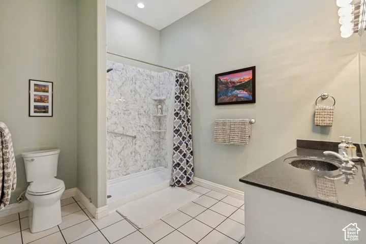 Tiled flooring, white toilet, shower with patterned wall surround, vanity with dark countertop and integrated sink, recessed lighting